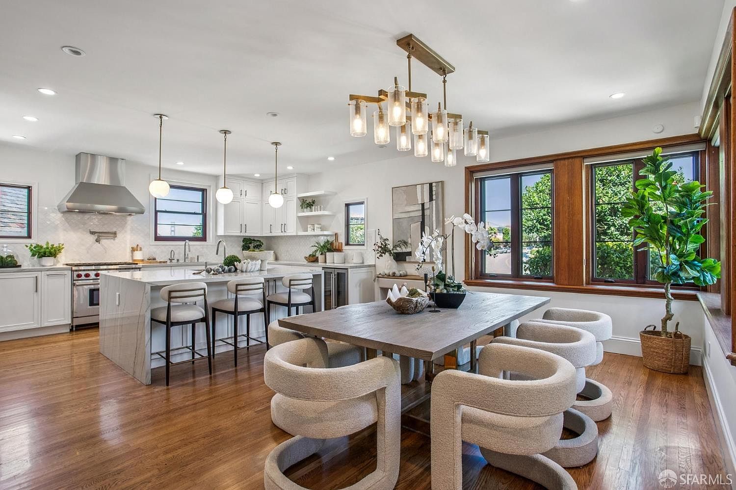 This open-concept space seamlessly blends a modern kitchen with a formal dining area, characterized by warm wood flooring and bright white cabinetry. The kitchen features a large island with bar seating, stainless steel appliances, and a sleek range hood, while the dining area is anchored by a substantial wooden table surrounded by contemporary, curved-back chairs. Large windows with dark frames allow natural light to flood the room, highlighting the elegant chandelier above the dining table and the lush greenery of the potted plant in the corner.