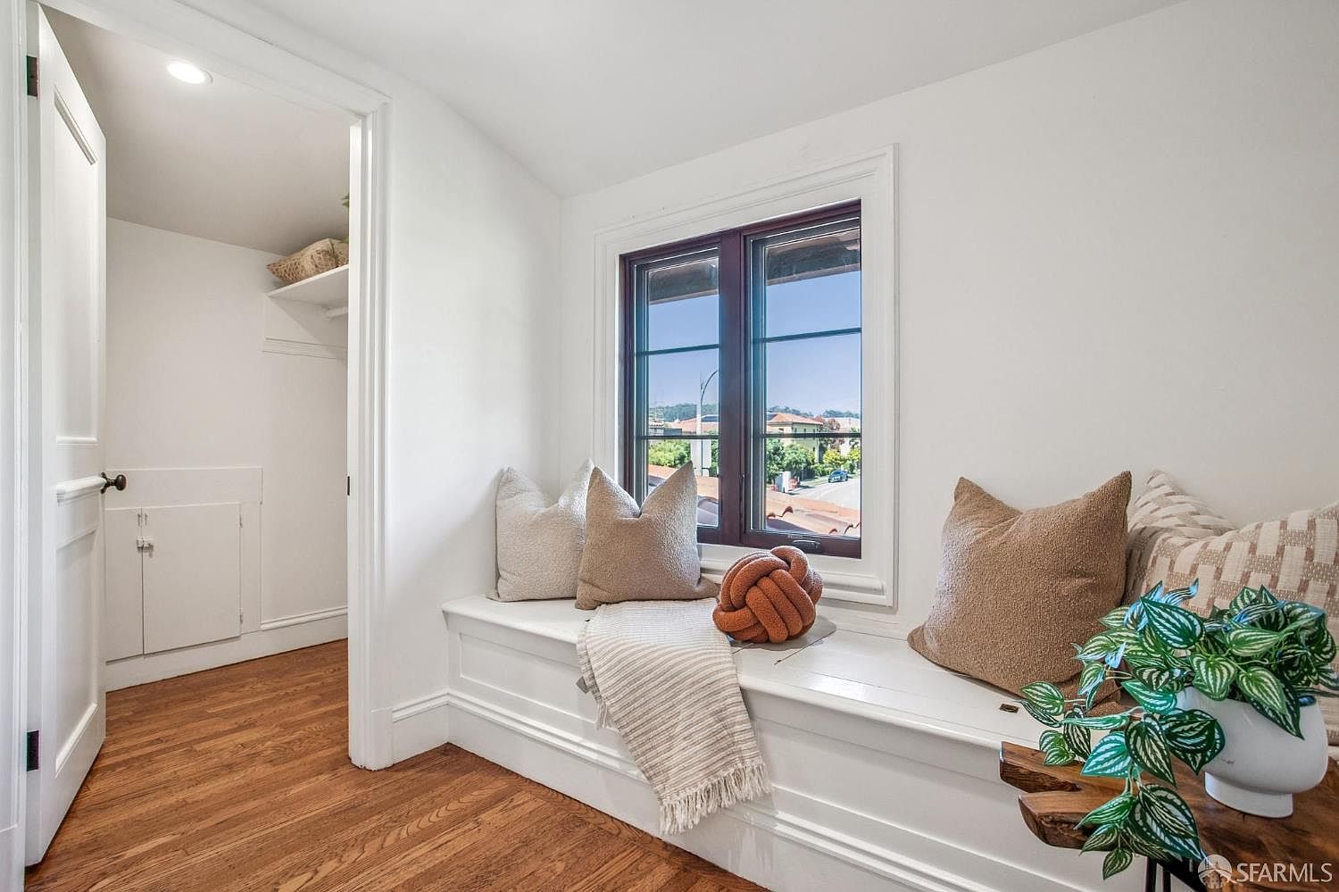 This cozy nook features a built-in window seat with plush decorative pillows and a throw blanket, creating a comfortable reading area. The space is bright and airy with white walls, light wood flooring, and a view through the window to the neighborhood outside. To the left, an open doorway leads into a small walk-in closet, adding functional storage to this charming transitional space.