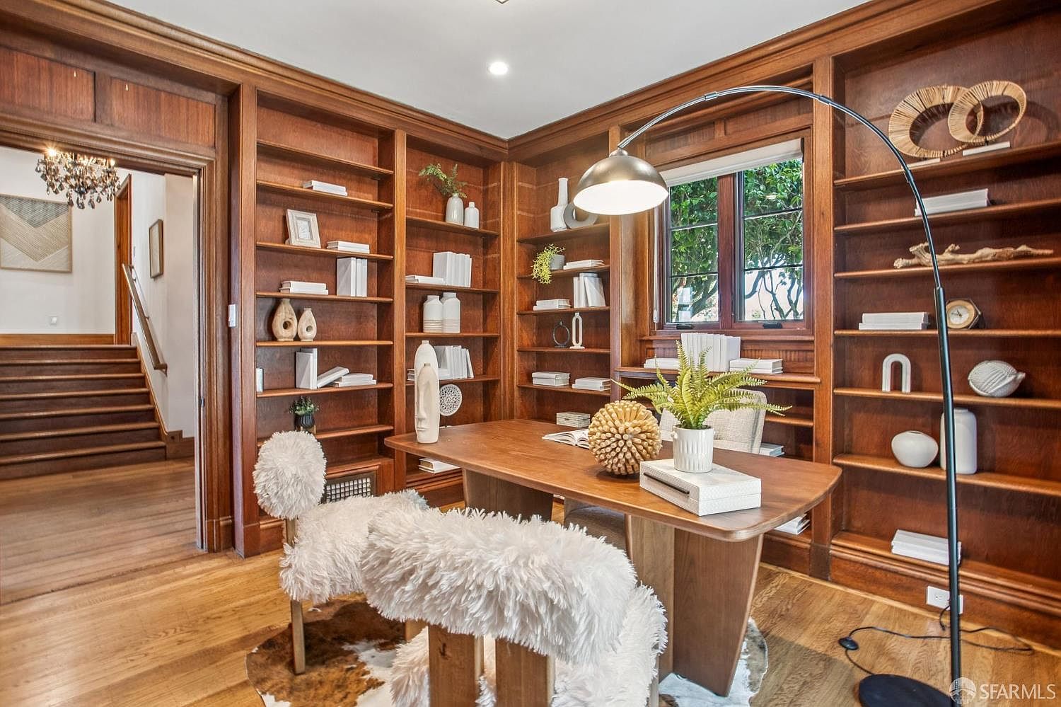 This sophisticated home office features extensive built-in wood shelving that spans the walls, creating a warm and scholarly atmosphere. A central wooden desk is paired with two unique, textured white chairs, while a large arched floor lamp provides focused lighting. The room is accented by a patterned rug and offers a view into an adjacent hallway with a staircase, emphasizing the home's traditional architectural character.