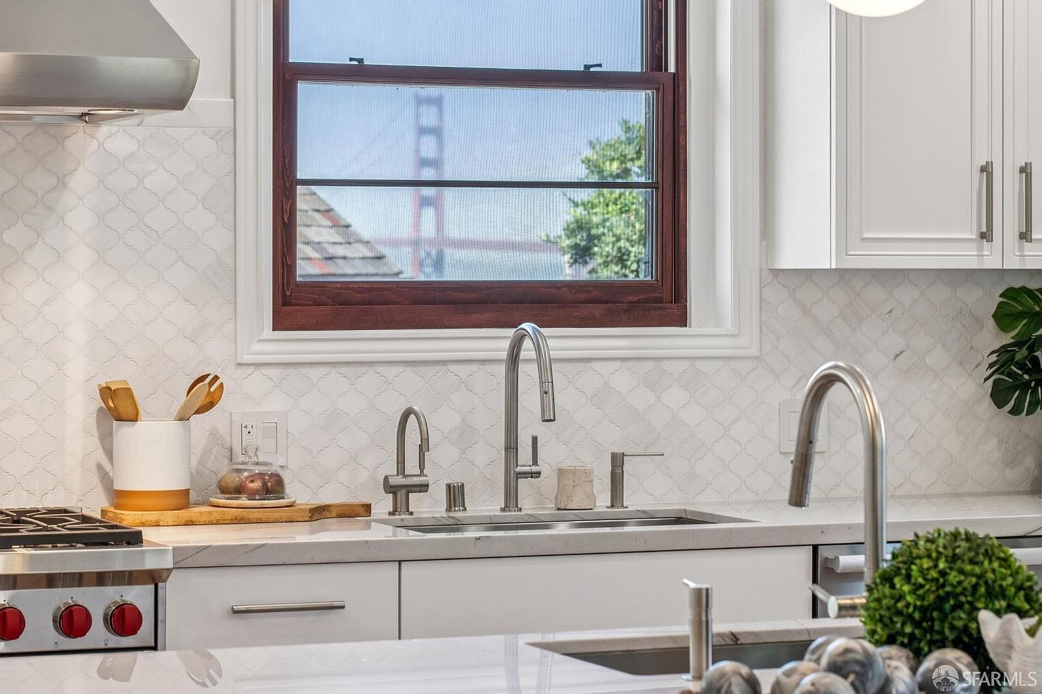 This bright, modern kitchen features white cabinetry and a patterned tile backsplash that adds texture to the space. A central window provides a unique, framed view of the Golden Gate Bridge, while stainless steel fixtures and a professional-style range highlight the high-end finishes. The perspective is a close-up, eye-level shot focusing on the sink area and the iconic bridge view beyond.