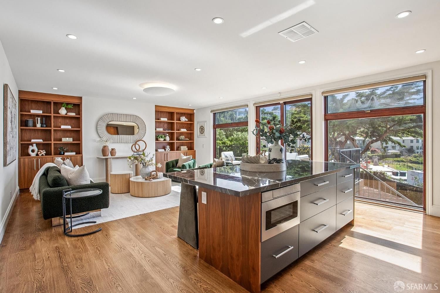 This open-concept living and kitchen area features warm wood flooring and a large kitchen island with a dark stone countertop and integrated microwave. The adjacent living space is anchored by built-in shelving units and a modern sofa, creating a cohesive and inviting atmosphere. Large windows provide ample natural light and views of the surrounding greenery, enhancing the bright and airy feel of the home.