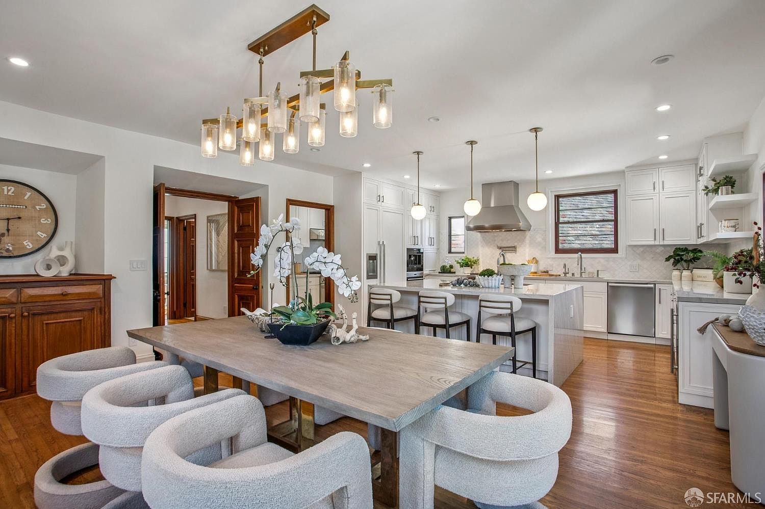 This open-concept space seamlessly blends a dining area with a modern kitchen, featuring a large wooden dining table surrounded by textured, curved chairs. The kitchen is anchored by a spacious island with bar seating, complemented by white cabinetry, stainless steel appliances, and a professional-grade range hood. The room is illuminated by a striking multi-light chandelier above the dining table and warm pendant lights over the island, creating an inviting and sophisticated atmosphere.