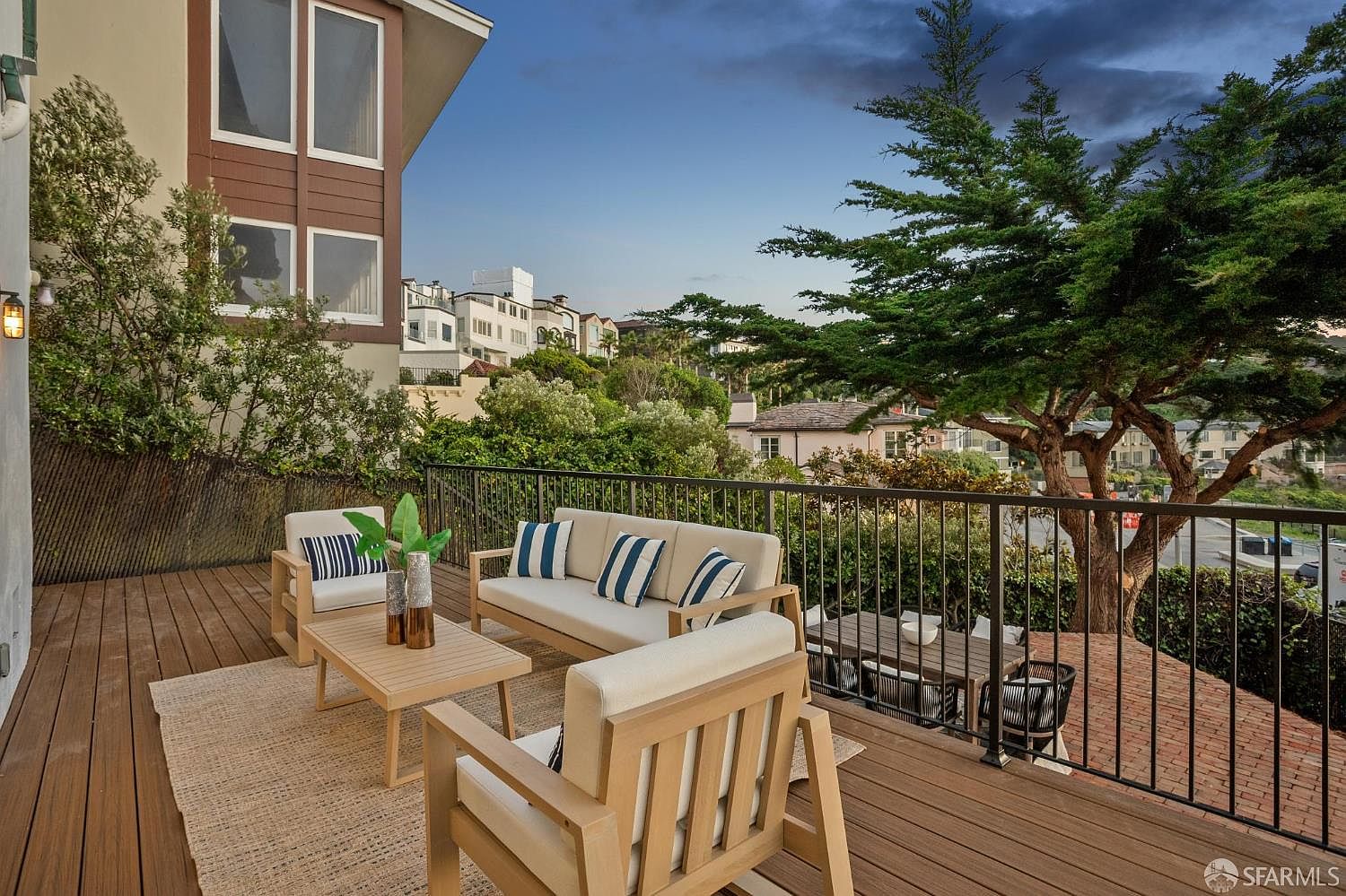This elevated wooden deck offers a comfortable outdoor living space furnished with a light-toned sofa, matching armchairs, and a central coffee table atop a textured rug. The deck is enclosed by a black metal railing that overlooks a lush, tree-filled landscape and neighboring homes in the distance. A separate dining area is visible on a lower brick patio level, creating a layered and inviting atmosphere for relaxation or entertaining.