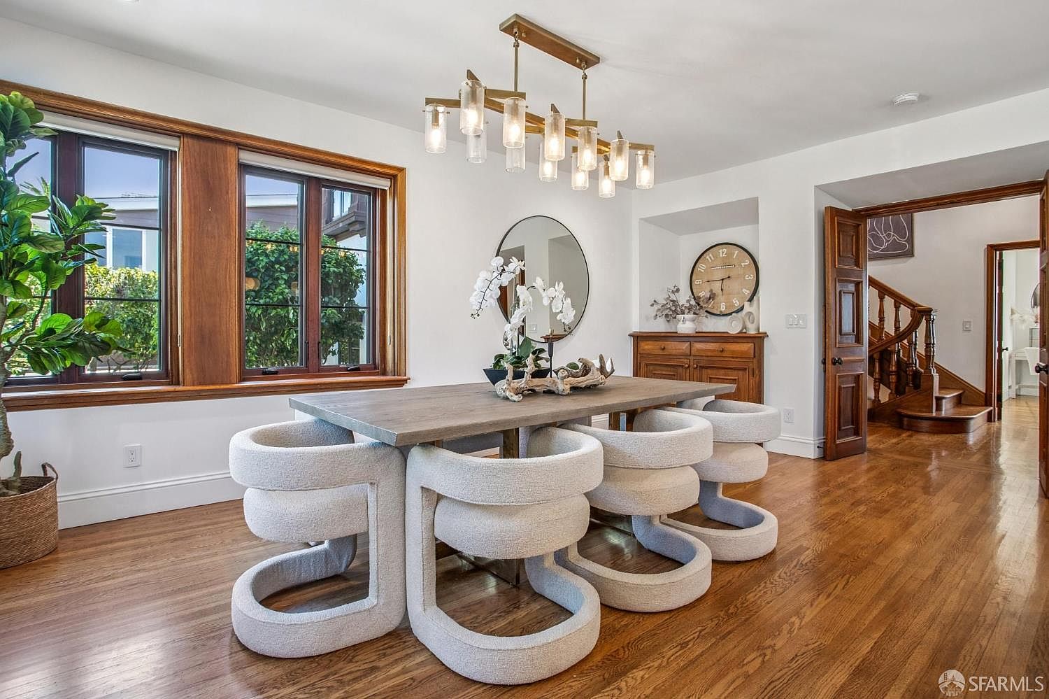 This elegant dining room features a contemporary wooden table surrounded by six unique, sculptural white upholstered chairs. A striking multi-light chandelier hangs above the table, while a large circular mirror and a wall-mounted clock add character to the space. The room is finished with warm wood flooring and classic wood-trimmed windows, creating a sophisticated and inviting atmosphere.