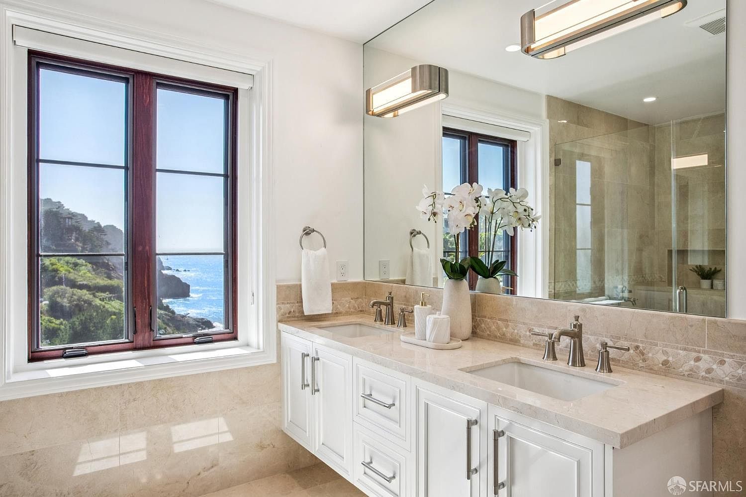 This bright and airy primary bathroom features a double vanity with white cabinetry and a light-toned stone countertop. A large mirror spans the wall above the sinks, reflecting the natural light and the scenic ocean view visible through the adjacent window. The space is finished with neutral stone wall tiles and modern light fixtures, creating a serene and luxurious atmosphere.