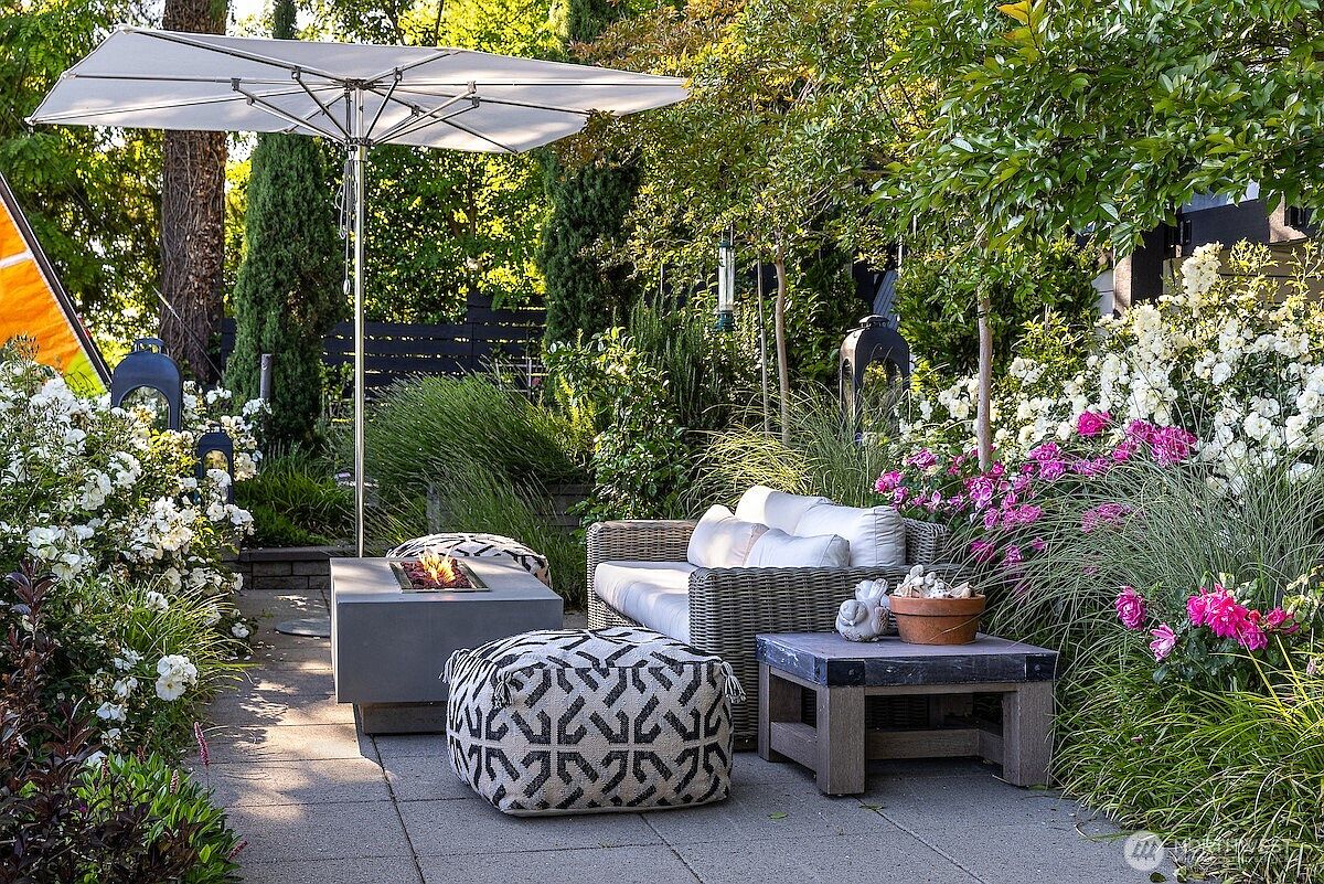 This inviting outdoor patio space features a modern gray wicker sofa with plush cushions, paired with two patterned ottomans and a sleek, low-profile fire pit table. A large, square cantilever umbrella provides shade over the seating area, which is surrounded by lush landscaping, including blooming white and pink roses and ornamental grasses. The scene is captured from a ground-level perspective, highlighting a serene and well-manicured garden retreat perfect for relaxation.
