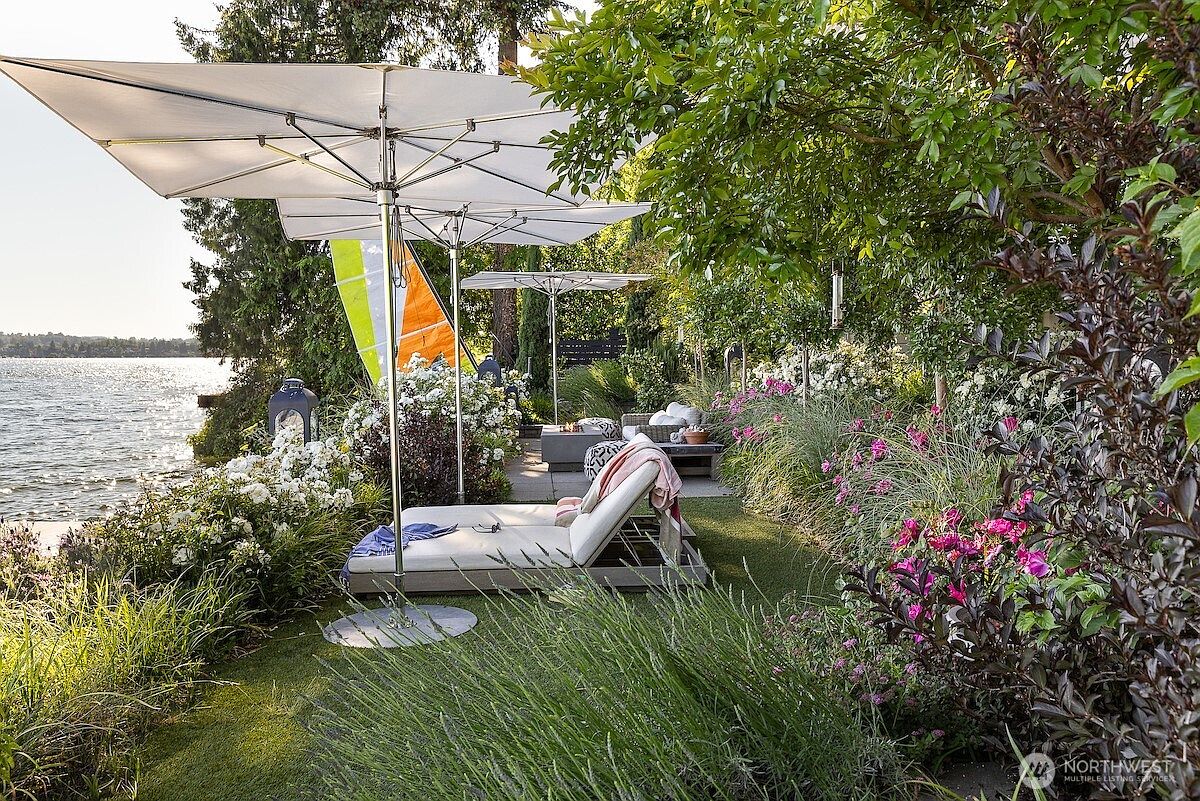 This serene waterfront patio area features a comfortable lounge chair shaded by a large, modern umbrella, overlooking a calm lake. The space is beautifully landscaped with lush greenery, vibrant pink flowers, and white rose bushes, creating a private and tranquil retreat. A stone-paved path leads toward a secondary seating area with a fire pit, emphasizing a sophisticated outdoor living space perfect for relaxation.
