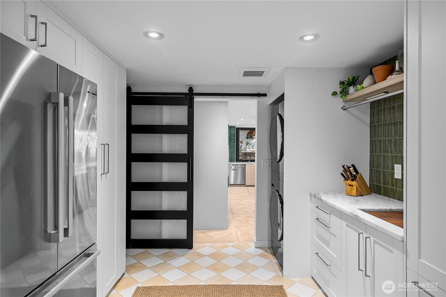 This functional laundry room features a stacked washer and dryer unit set against a light-colored wall with a green tiled backsplash and a stone countertop. A sliding barn door with frosted glass panels provides a stylish transition to the adjacent kitchen area, which is visible in the background. The space is finished with a light-toned, diamond-patterned tile floor and recessed lighting, creating a clean and organized utility area.