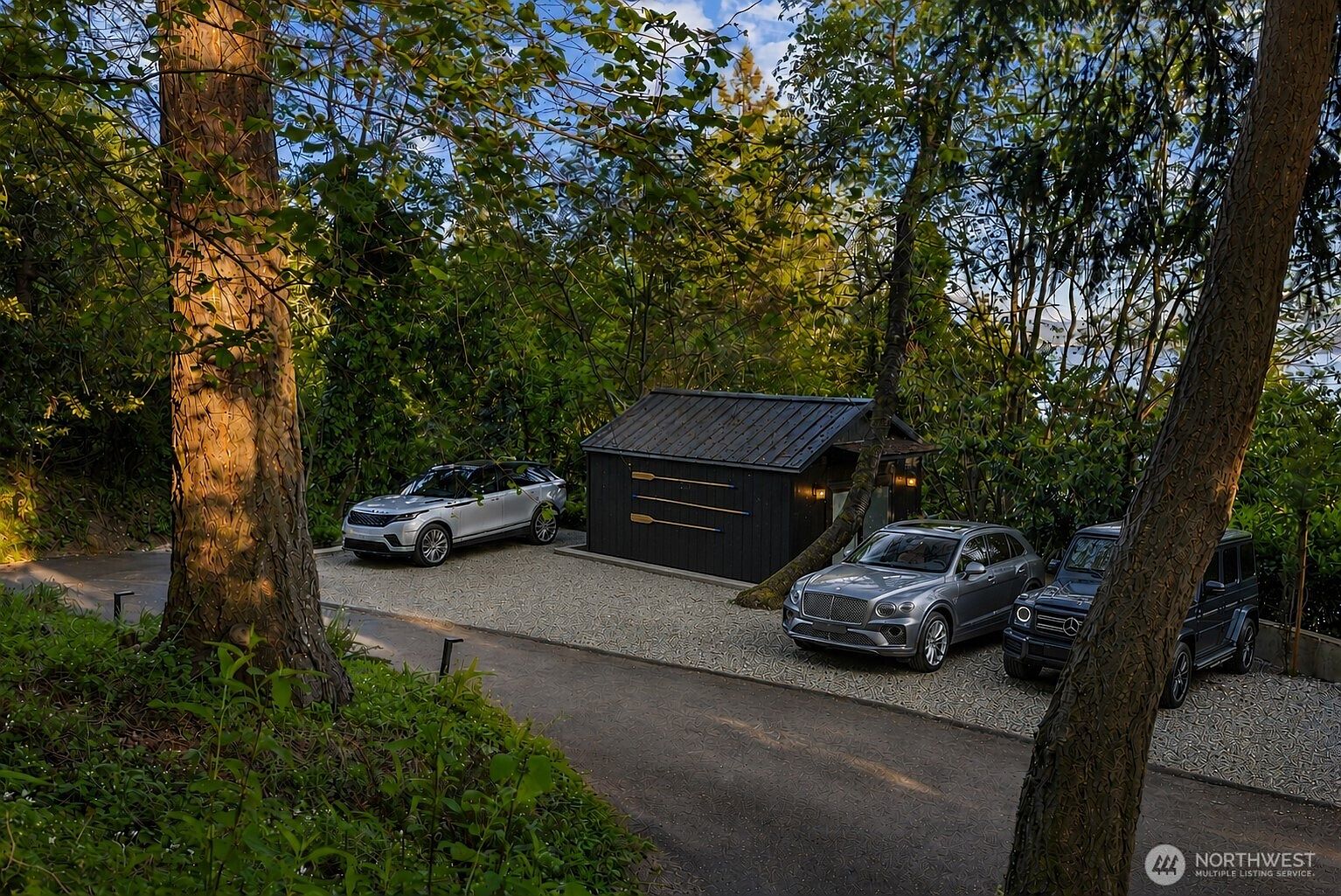 This outdoor scene features a gravel parking area nestled among mature trees, showcasing a small, dark-sided shed adorned with decorative oars. Three luxury vehicles are parked in the space, creating a sophisticated and private atmosphere. The perspective is slightly elevated, capturing the lush, wooded surroundings and the serene, secluded nature of the property.