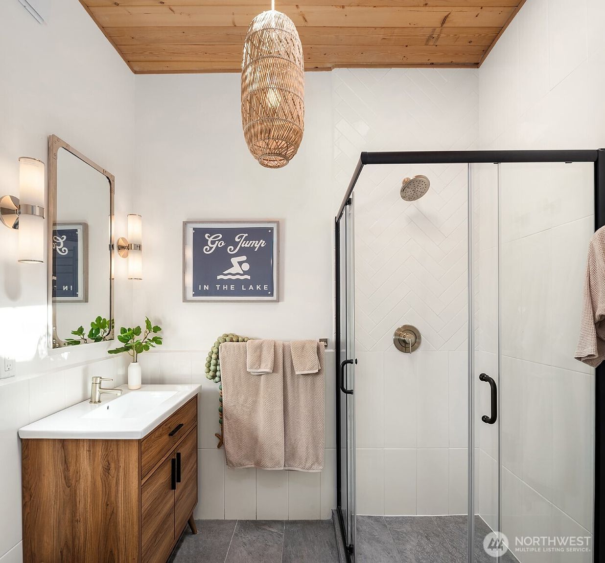 This modern bathroom features a wood-toned vanity with a white sink, complemented by a large rectangular mirror and dual wall-mounted light fixtures. A glass-enclosed shower with a herringbone-patterned tile surround and a rainfall showerhead serves as a focal point, while a woven pendant light adds a touch of organic texture. The space is finished with a playful 'Go Jump in the Lake' wall sign, creating a charming, vacation-inspired aesthetic.