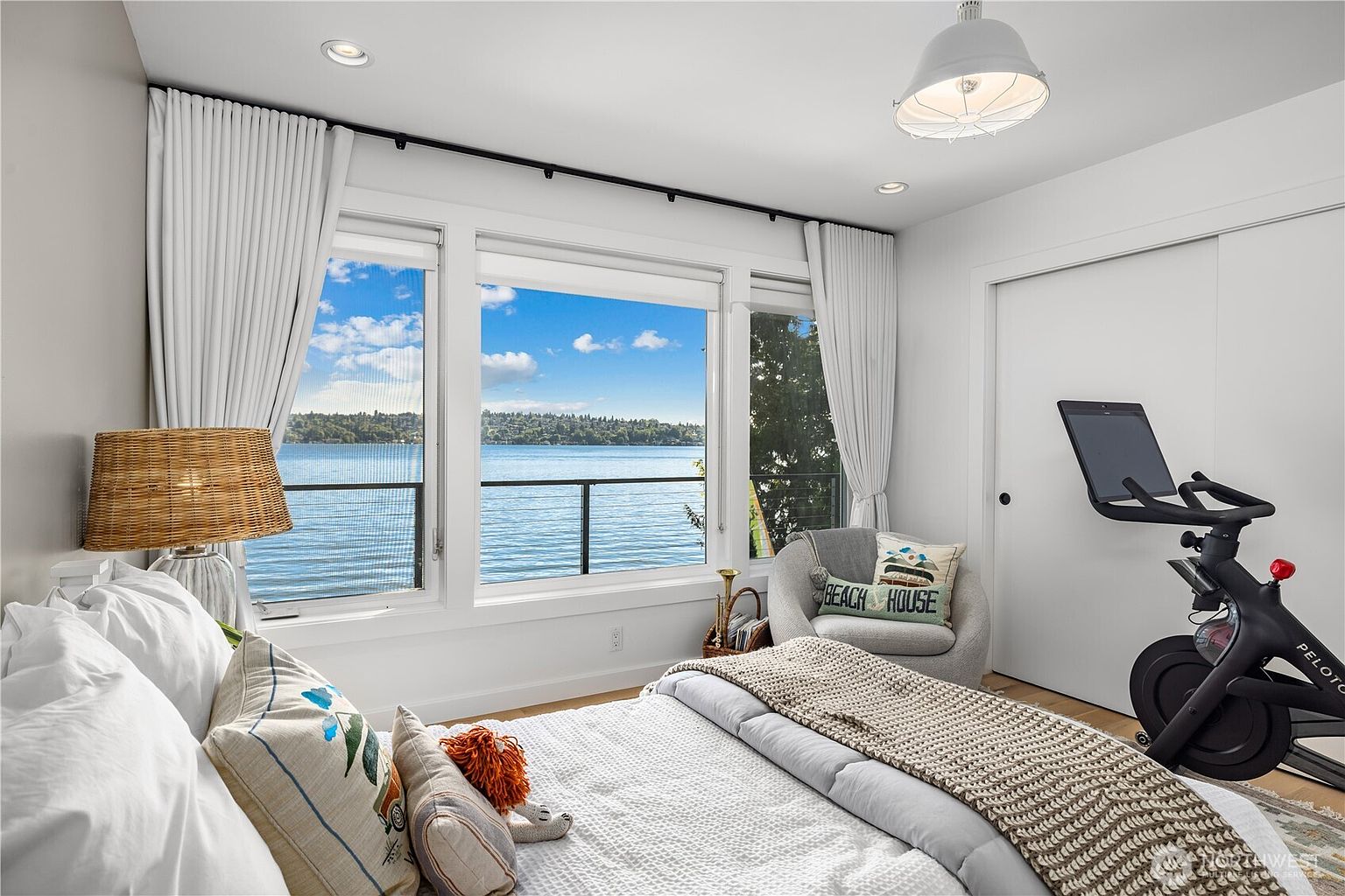 This bright and airy bedroom features a comfortable bed with layered bedding and a textured throw blanket, positioned to face a set of large windows that provide a stunning, unobstructed view of the lake. To the side, a cozy armchair with a decorative pillow sits near a stationary exercise bike, creating a multi-functional space. The room is illuminated by a modern pendant light and a bedside lamp with a woven shade, contributing to a serene and inviting atmosphere.
