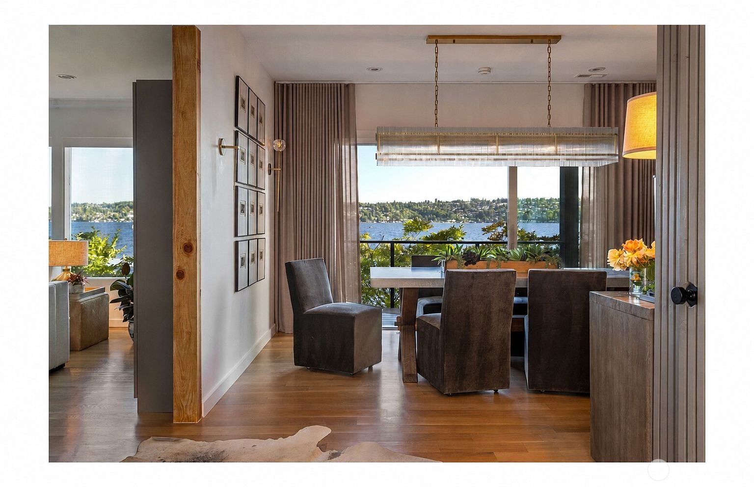 This elegant dining area features a large rectangular table surrounded by upholstered chairs, positioned to take full advantage of the expansive water view through large windows. A sophisticated, linear glass-and-metal chandelier hangs above the table, while a gallery wall of framed art adds a personal touch to the space. The room flows seamlessly into an adjacent living area, with warm wood flooring and a natural, neutral color palette creating a serene and inviting atmosphere.
