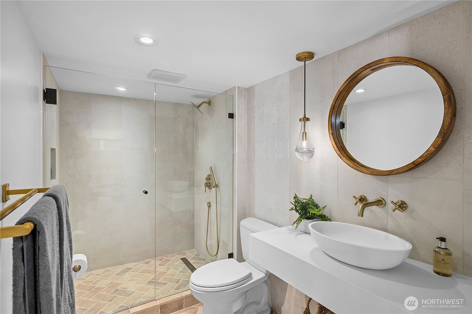 This modern bathroom features a floating white vanity with a vessel sink and gold-toned wall-mounted fixtures. A glass-enclosed walk-in shower showcases patterned tile flooring in a herringbone layout, complemented by neutral wall tiles. A round mirror with a warm wood frame hangs above the vanity, illuminated by a single pendant light, creating a clean and sophisticated aesthetic.