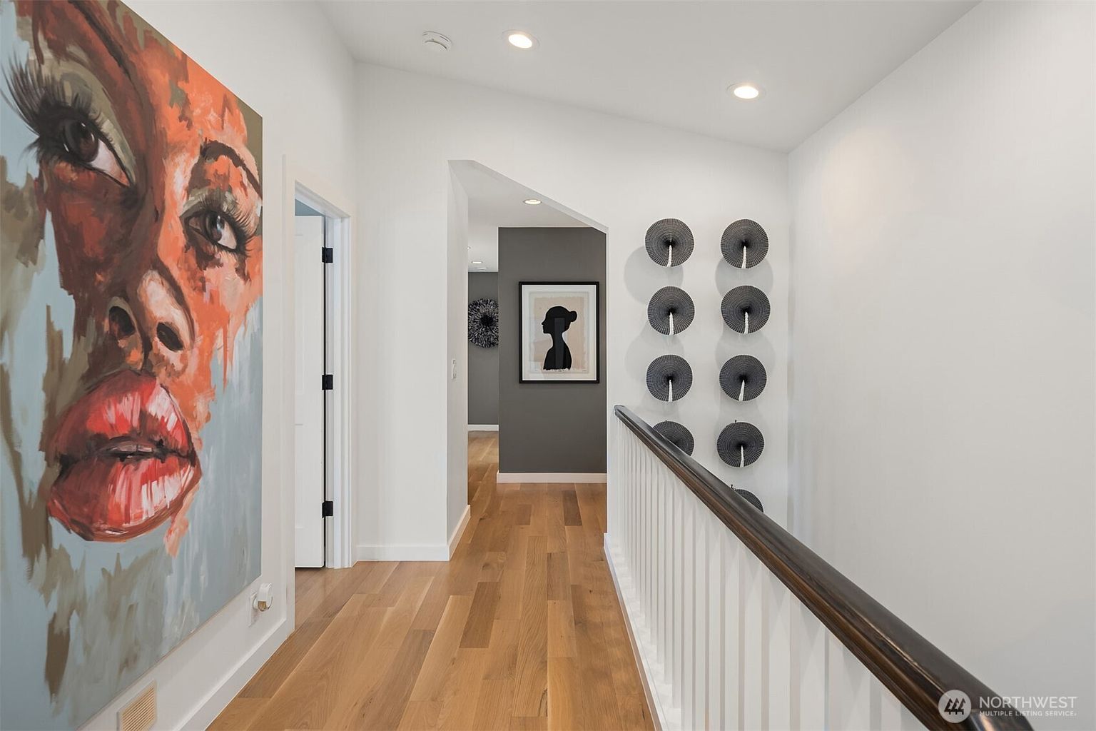 This bright hallway features light wood flooring and a white railing overlooking a staircase. The walls are adorned with a large, vibrant portrait painting on the left and a series of circular decorative wall art pieces on the right. A doorway leads into a secondary space with a dark accent wall and framed artwork, creating a modern and artistic aesthetic.