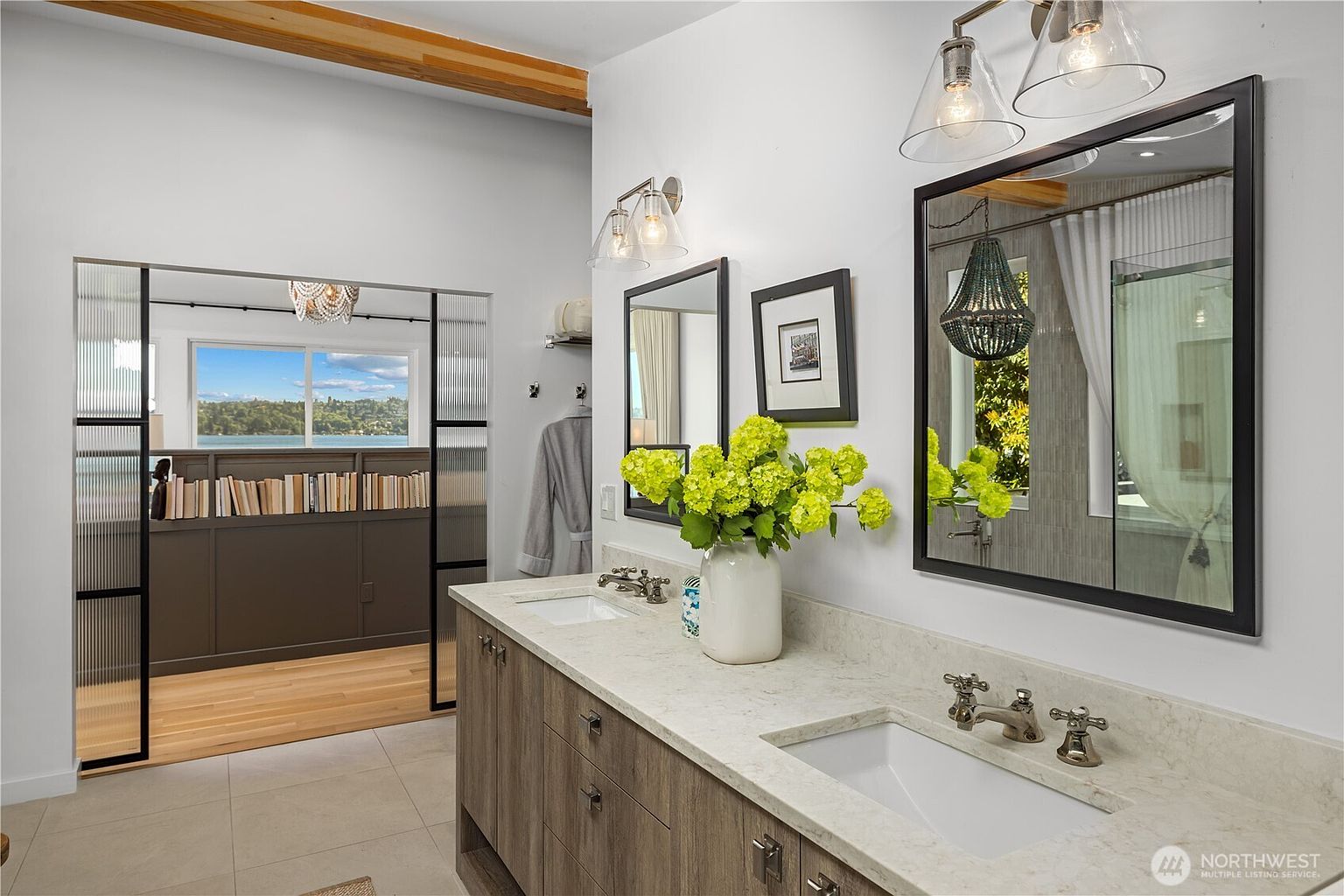 This bright and modern primary bathroom features a dual-sink vanity with light wood cabinetry and a neutral stone countertop. A large, black-framed mirror hangs above the vanity, reflecting a view of a chandelier and a glass-enclosed shower area. To the left, a sliding glass door with a metal frame opens into an adjacent room, which offers a scenic view of the water through large windows.