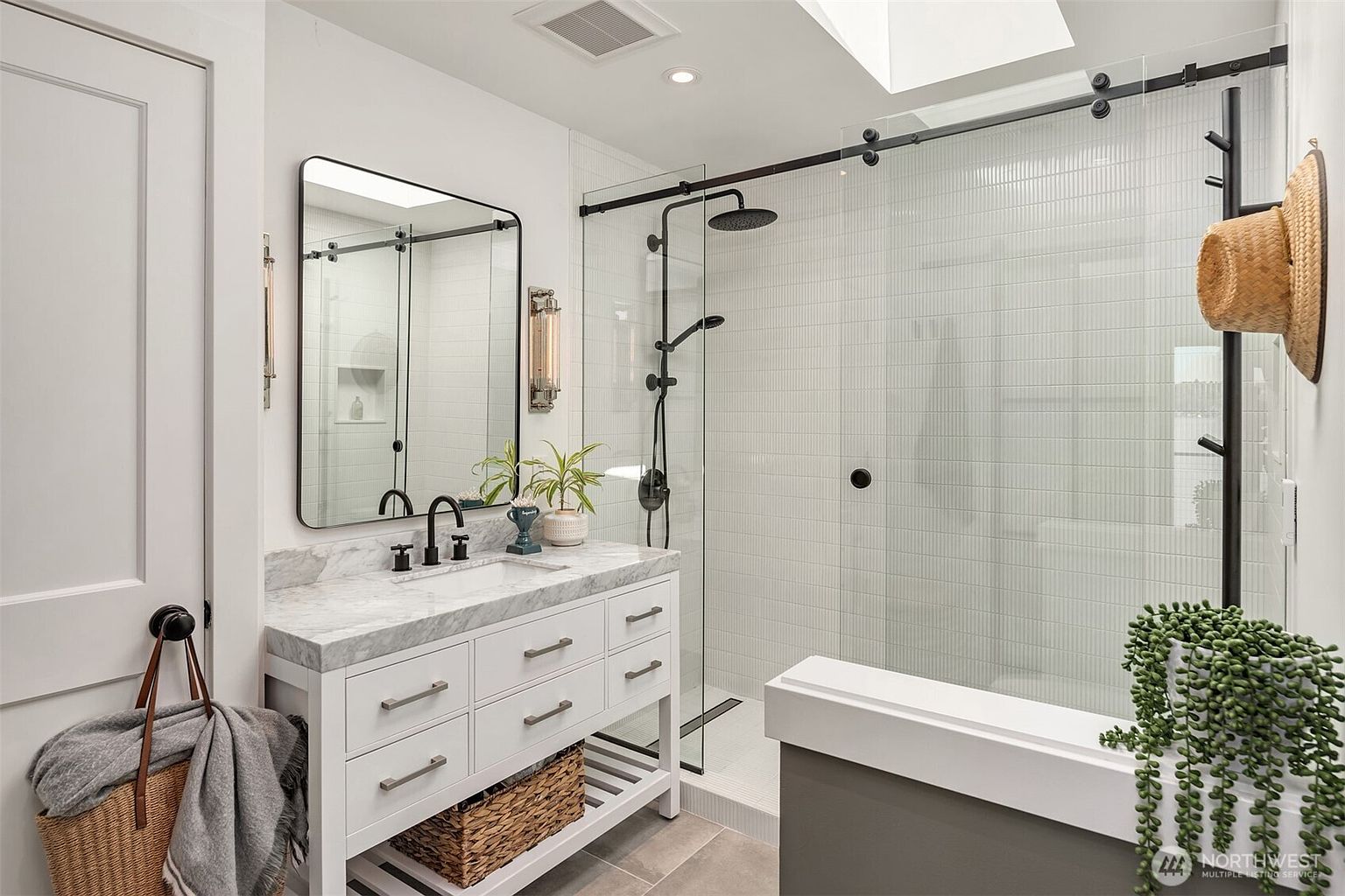 This modern bathroom features a white vanity with a stone countertop, a rectangular mirror with a black frame, and a walk-in shower with a glass sliding door and black hardware. The shower is tiled with white vertical subway tiles and includes both a rainfall showerhead and a handheld sprayer. A woven basket sits on the lower shelf of the vanity, and a straw hat hangs on the wall, adding a touch of casual, coastal-inspired decor to the clean, bright space.