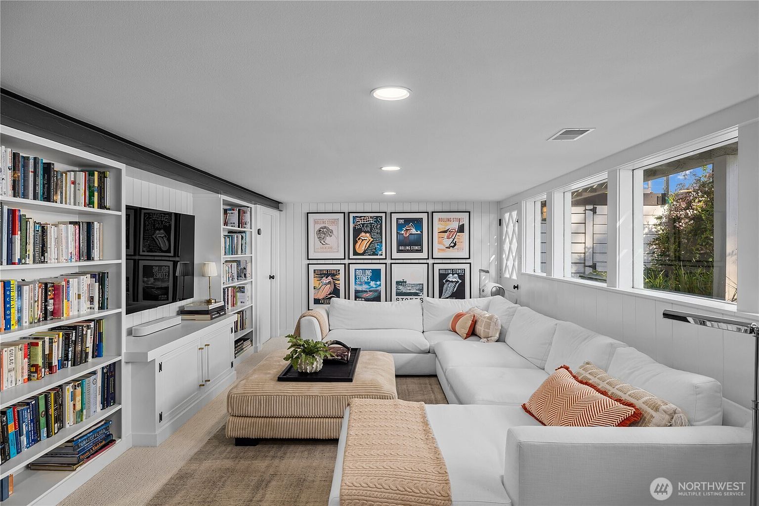 This cozy living room features a large, white L-shaped sectional sofa paired with a textured, neutral-toned ottoman. The space is anchored by built-in white shelving units flanking a wall-mounted television, while a gallery wall of Rolling Stones posters adds a vibrant, personal touch. Large windows along the right wall allow for ample natural light, creating a bright and inviting atmosphere.