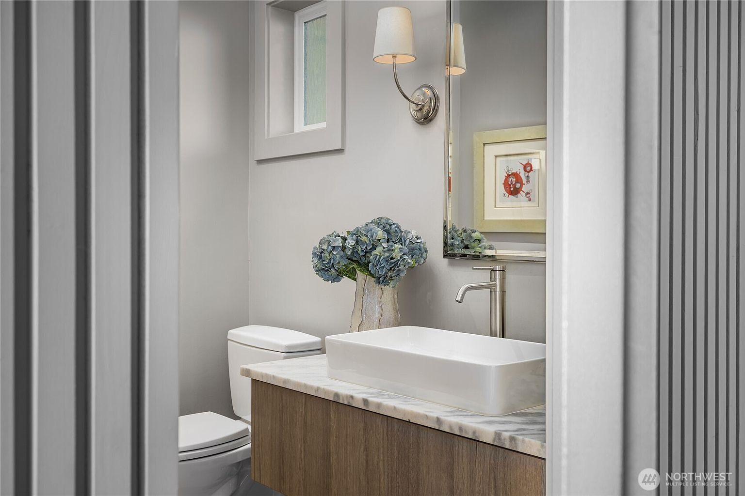 This compact powder room features a modern vessel sink set atop a light wood vanity with a stone countertop. A wall-mounted light fixture illuminates the space, which includes a toilet and a framed piece of art on the wall. The neutral color palette and clean lines create a polished, contemporary aesthetic.