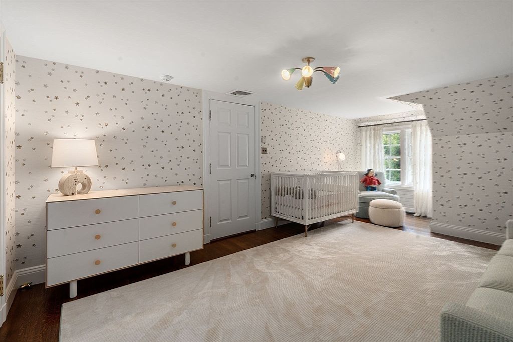 This charming nursery features whimsical star-patterned wallpaper and a light-toned wood dresser topped with a decorative lamp. A white crib is positioned near a window with soft curtains, while a plush armchair and ottoman create a cozy seating area. The room is finished with a large, neutral-toned area rug over dark wood flooring, providing a warm and inviting atmosphere.