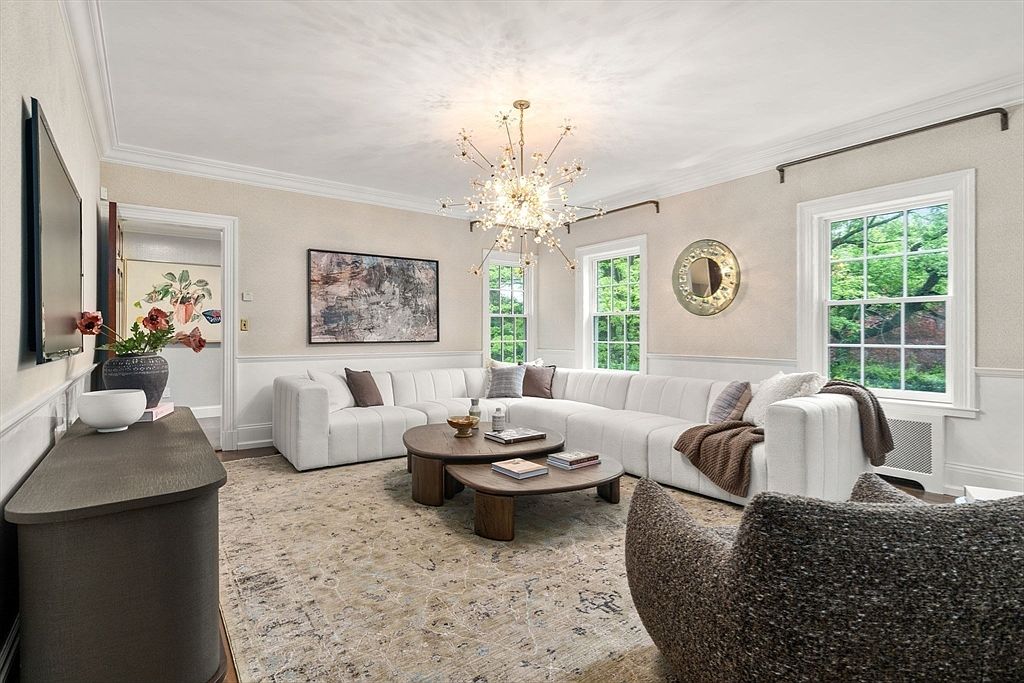 This elegant living room features a large, white sectional sofa arranged around two circular wooden coffee tables, set atop a textured, neutral-toned area rug. A striking, multi-armed starburst chandelier serves as the focal point, while tall windows with white trim allow natural light to brighten the space. The room is decorated with a large abstract wall art piece, a circular wall mirror, and a dark console table, creating a sophisticated and inviting atmosphere.