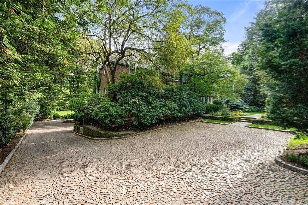 This image captures a side view of a traditional brick residence nestled within a lush, mature landscape. A wide, circular driveway paved with cobblestone-style pavers leads toward the house, which is partially obscured by dense, well-maintained shrubbery and tall, leafy trees. The perspective is from the driveway, emphasizing the secluded and private nature of the property's grounds.