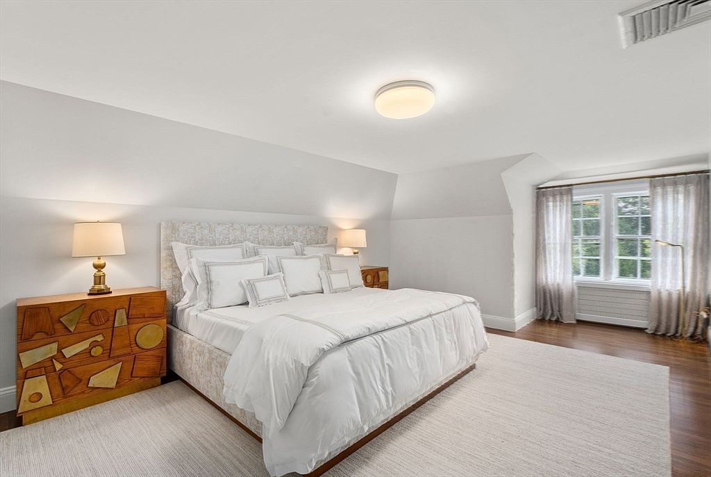 This spacious primary bedroom features a neutral color palette with a large, upholstered bed as the focal point, flanked by two bedside lamps. To the left, a unique wooden dresser with geometric, raised-relief detailing adds a touch of artistic texture to the room. The space is illuminated by a central flush-mount light fixture and natural light streaming through a set of windows dressed in sheer curtains, all set against a backdrop of hardwood floors and a large area rug.