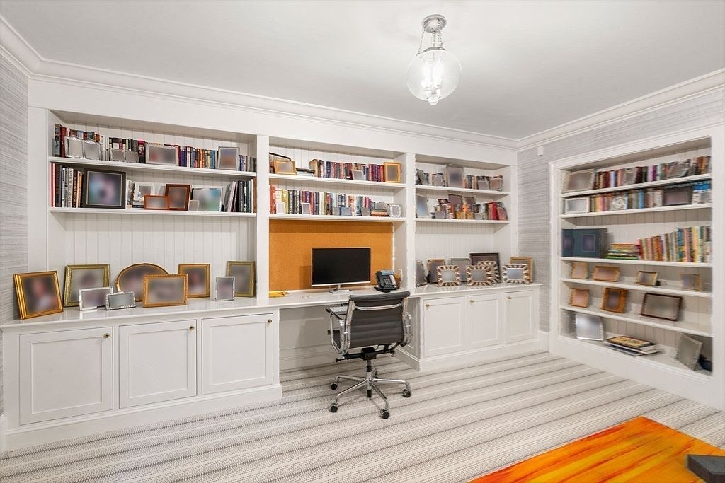 This organized home office features extensive built-in white cabinetry and shelving that spans the entire wall, providing ample storage and display space. A central desk area with a computer monitor and office chair is set against a warm-toned cork board, while the room is finished with neutral striped carpeting and a classic glass pendant light. The space offers a clean, professional aesthetic with a symmetrical layout that maximizes functionality.