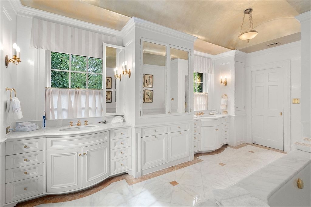This elegant primary bathroom features a symmetrical layout with dual vanities, white cabinetry, and light-toned stone countertops. A central storage unit with mirrored doors sits between the sinks, while a vaulted ceiling with a warm, metallic finish adds a touch of luxury. The space is brightened by large windows with soft drapery and classic wall sconces, creating a refined and airy atmosphere.