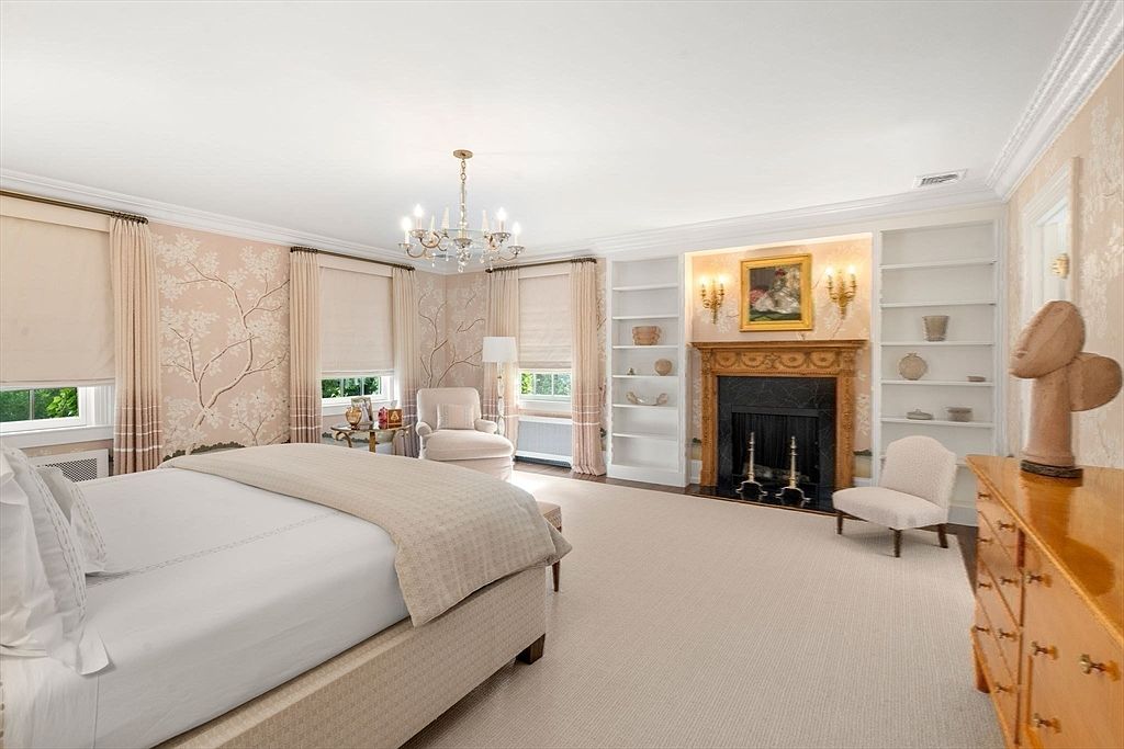 This elegant primary bedroom features soft, neutral tones and sophisticated wallpaper with a delicate tree motif. The room is anchored by a large bed with a textured headboard, complemented by a fireplace with a dark stone surround and a wooden mantel flanked by built-in shelving. Large windows with light-filtering shades allow for ample natural light, creating a serene and luxurious atmosphere.