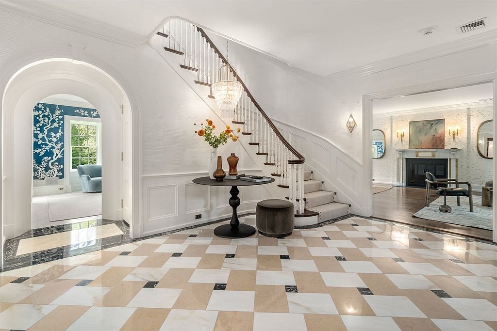 This elegant foyer features a grand staircase with a white balustrade and a runner, set against a backdrop of crisp white walls and wainscoting. The floor is finished in a classic checkerboard pattern of light and dark stone tiles, leading the eye toward an arched entryway on the left and a spacious living area on the right. A crystal chandelier hangs above a circular pedestal table, creating a sophisticated and welcoming first impression.