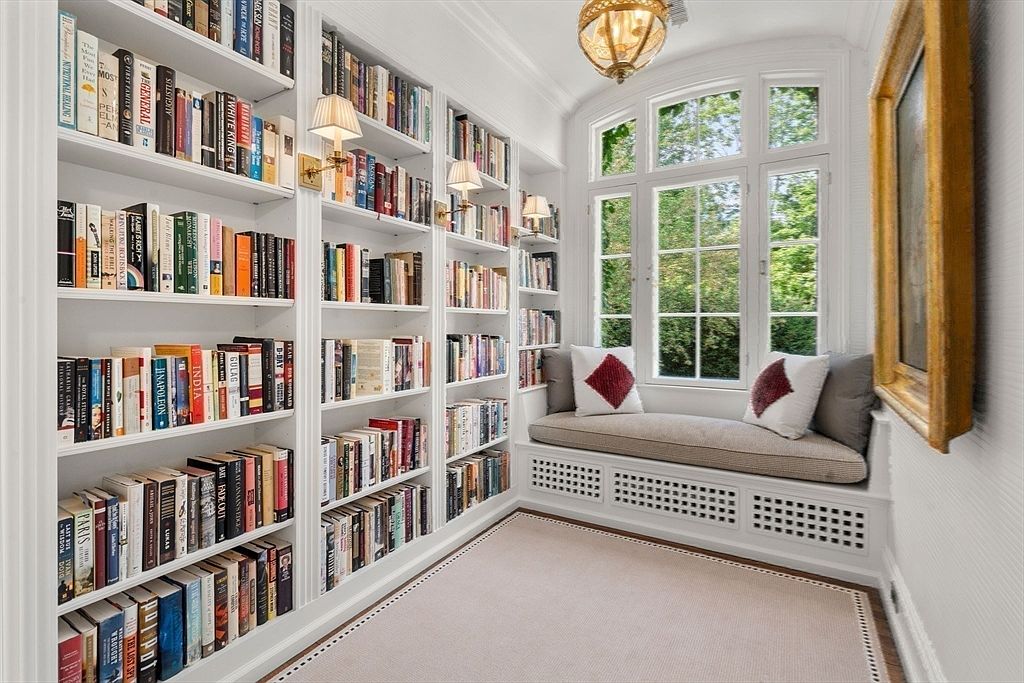 This cozy, light-filled reading nook or study features extensive built-in white bookshelves lining the walls, filled with a diverse collection of books. A comfortable window seat with plush cushions and decorative pillows sits beneath a large, multi-pane window that overlooks lush greenery. The space is accented by warm wall-mounted lighting and a classic chandelier, creating an inviting and scholarly atmosphere.
