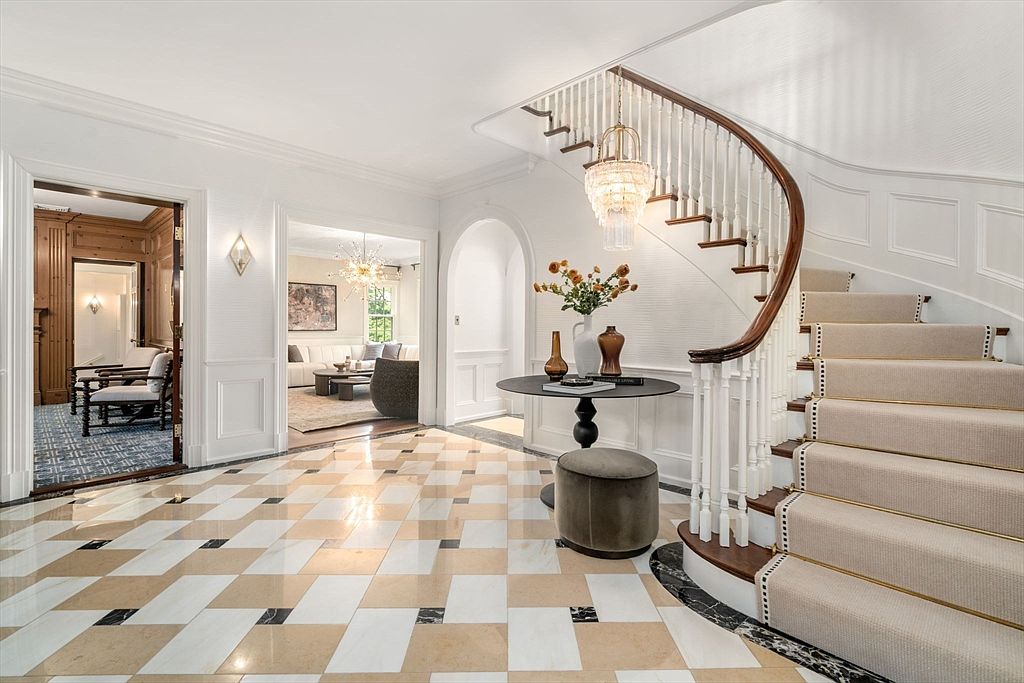 This elegant foyer features a grand curved staircase with a dark wood handrail and a neutral carpet runner. The space is anchored by a checkered stone floor and a central pedestal table topped with a floral arrangement, illuminated by a crystal chandelier. Through the arched openings, one can see glimpses of a wood-paneled study and a bright, airy living area, creating a sophisticated and welcoming first impression.