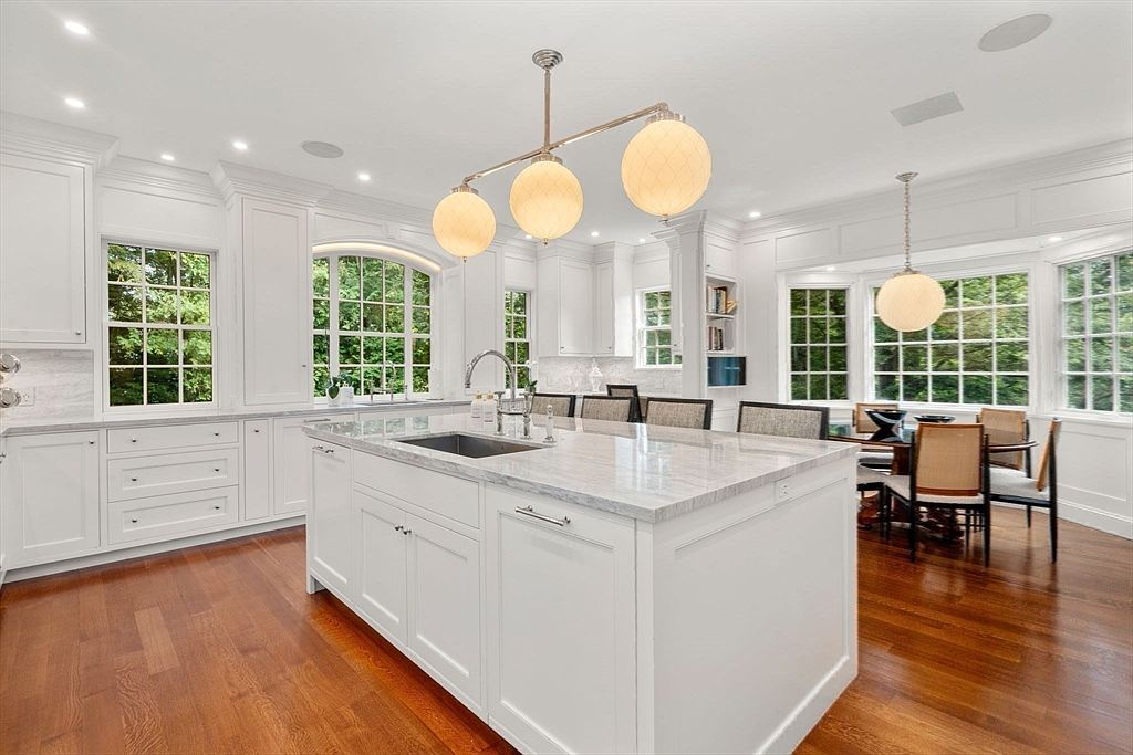 This bright, spacious kitchen features white cabinetry, a large central island with a stone countertop, and warm-toned wood flooring. A three-globe pendant light fixture hangs above the island, while a separate dining area with a round table and chairs is visible to the right. Large windows throughout the space provide ample natural light and views of the surrounding greenery, creating an airy and inviting atmosphere.