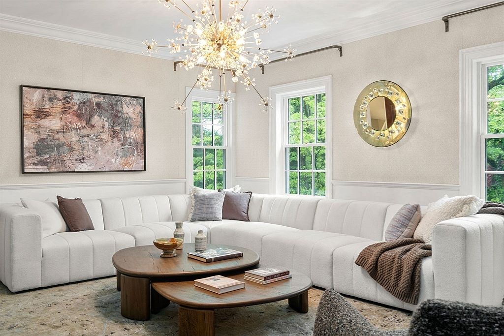 This bright and inviting living room features a large, white, channel-tufted sectional sofa arranged in an L-shape, providing ample seating. The space is centered around a pair of nested, round wooden coffee tables and illuminated by a striking, multi-armed starburst chandelier. Large windows with views of lush greenery and a circular decorative mirror on the wall enhance the airy, sophisticated atmosphere.
