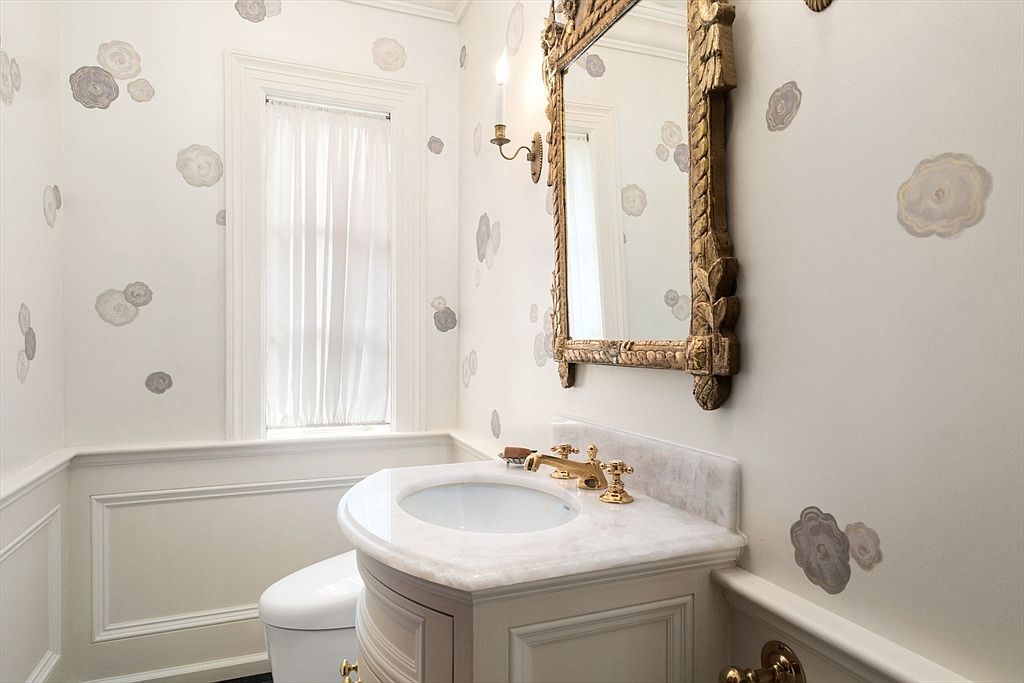 This elegant powder room features a vanity with a light-toned stone countertop and a gold-toned faucet. The walls are adorned with a whimsical floral decal pattern, complemented by a decorative, ornate gold-framed mirror and a matching wall sconce. A window with sheer curtains provides soft natural light, while the lower walls are finished with classic wainscoting.
