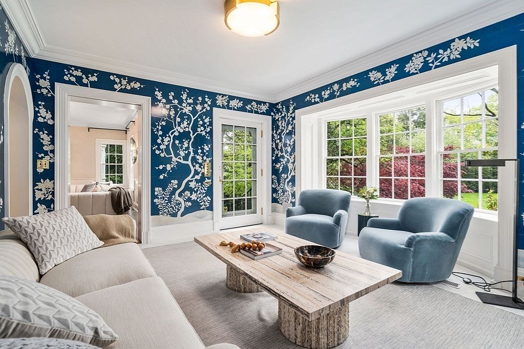 This elegant living room features striking blue wallpaper with a white botanical pattern, creating a sophisticated and artistic atmosphere. The space is furnished with a neutral-toned sofa and two plush blue armchairs arranged around a unique, textured stone coffee table. Large windows allow ample natural light to fill the room, offering a view of the lush greenery outside.