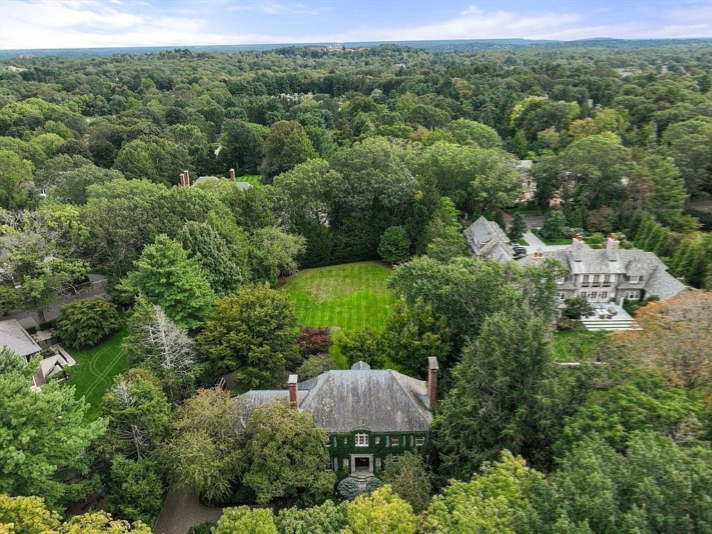 This high-angle aerial perspective captures a sprawling residential property nestled within a dense, lush forest. The scene features a large, traditional-style home with a dark shingled roof, surrounded by mature trees and a manicured central lawn. A second, larger residence is visible to the right, showcasing the expansive and private nature of this wooded neighborhood.