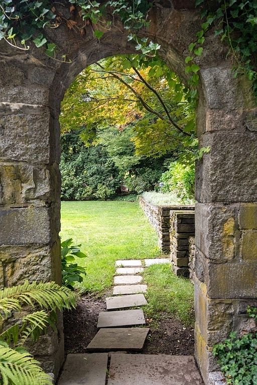 A rustic stone archway frames a view into a lush, private garden, creating a picturesque transition from a shaded area to a sunlit lawn. A path of rectangular stone pavers leads through the opening, flanked by a fern and climbing ivy, while a low stone retaining wall defines the edge of the green space. The scene evokes a sense of tranquility and timeless charm, highlighting the property's well-maintained landscaping.