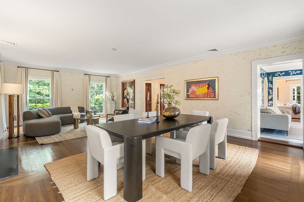 This elegant dining room features a dark, substantial dining table surrounded by six modern, light-colored upholstered chairs set atop a large textured area rug. The room is adorned with textured wallpaper, a framed piece of art, and a decorative vase centerpiece, creating a sophisticated and inviting atmosphere. A wide doorway leads into an adjacent living area, showcasing an open-concept layout with hardwood floors throughout.