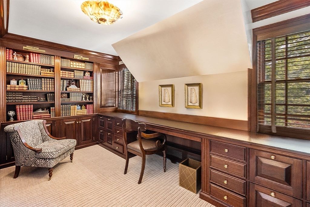 This sophisticated home office features extensive custom wood cabinetry and built-in bookshelves that span the wall, providing ample storage and display space. A long, integrated desk runs along the wall beneath a sloped ceiling, complemented by a patterned armchair and a classic wooden desk chair. The room is finished with neutral carpeting and warm, ambient lighting from a decorative ceiling fixture, creating a quiet and professional atmosphere.