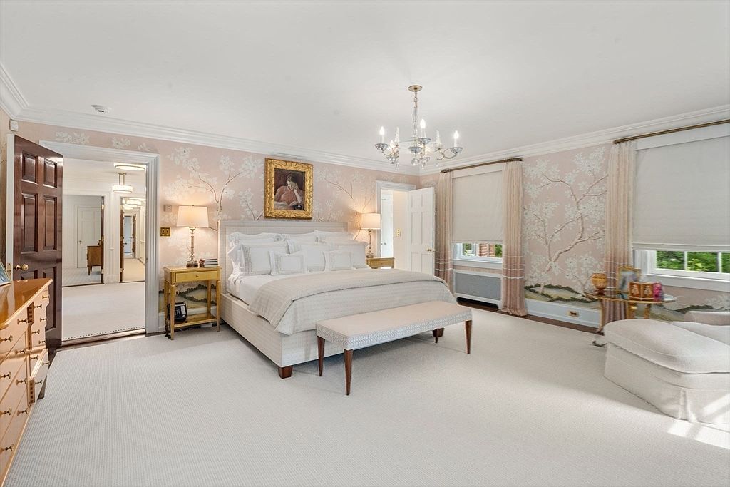 This elegant primary bedroom features soft pink floral wallpaper, a plush king-sized bed with a neutral upholstered headboard, and a matching bench at the foot. A crystal chandelier hangs from the center of the room, while large windows with light-filtering shades provide ample natural light. The space is framed by a dark wood door leading to a hallway and includes a comfortable armchair in the corner, creating a serene and sophisticated atmosphere.