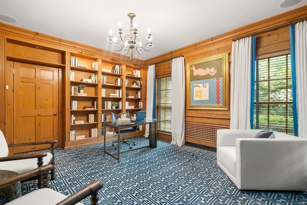This sophisticated home office features extensive light wood built-in shelving that spans the wall, providing ample storage and display space. A glass-topped desk sits centrally on a bold, geometric-patterned blue and white carpet, while a modern white armchair and a chandelier add a touch of elegance. The room is framed by tall windows with white curtains and blue trim, creating a bright and professional atmosphere.