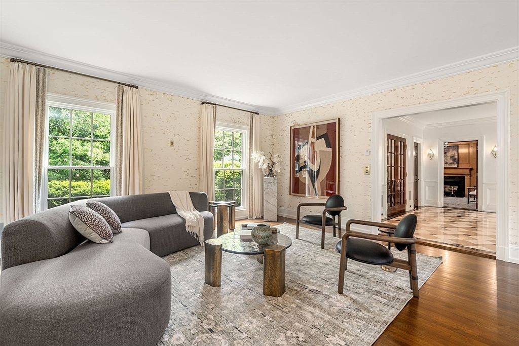 This elegant living room features a curved grey sofa, two modern armchairs, and a glass-topped coffee table with a metallic base, all arranged on a textured area rug. The space is brightened by large windows dressed in floor-length curtains and adorned with textured wallpaper, while a large abstract painting serves as a focal point on the wall. A wide doorway leads into a marble-floored hallway, providing a glimpse of a fireplace in the adjacent room, creating a sophisticated and open atmosphere.
