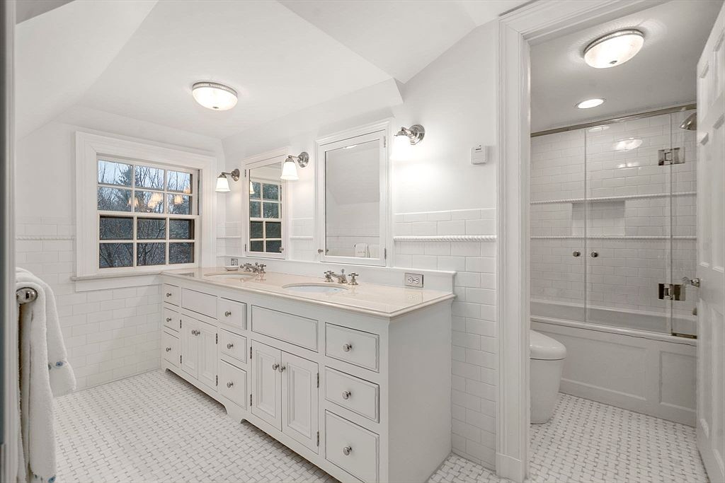 This bright and airy primary bathroom features a double vanity with a light-toned countertop, white cabinetry, and dual mirrors. The space is finished with white subway tile wainscoting and a classic basketweave-patterned tile floor, creating a clean, timeless aesthetic. A glass-enclosed shower and tub combination is visible through an open doorway, while a window above the vanity provides natural light.