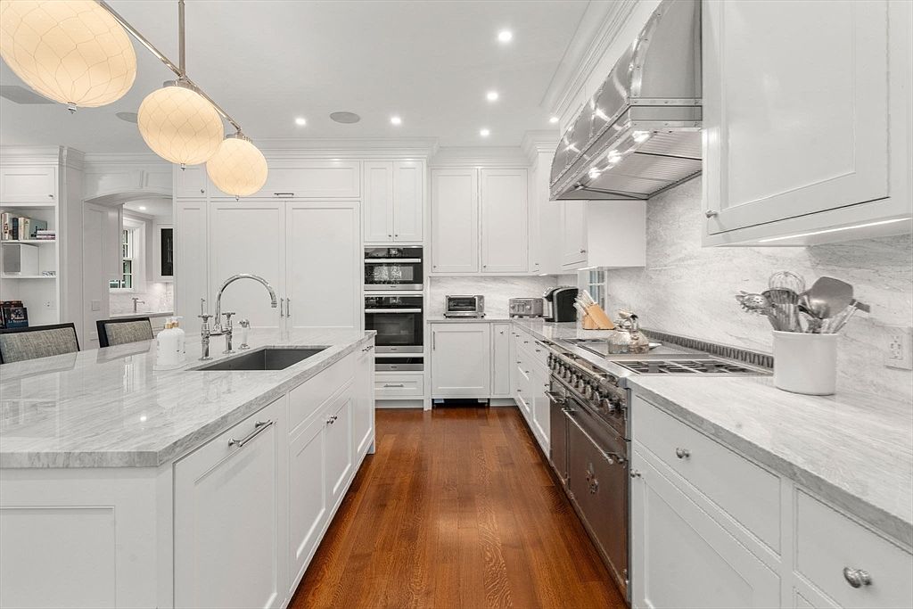 This bright and spacious kitchen features white cabinetry, a large central island with a stone countertop, and warm wood flooring. A professional-grade range with a stainless steel hood serves as a focal point, complemented by high-end appliances and elegant pendant lighting. The clean, classic design creates an inviting and functional space for cooking and entertaining.