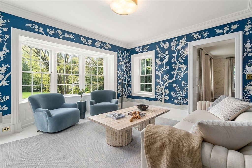 This elegant living room features striking blue wallpaper with a white botanical pattern, creating a sophisticated and cohesive aesthetic. The space is furnished with two plush blue armchairs and a light-toned sofa, centered around a textured, light-colored coffee table. Large windows allow ample natural light to fill the room, offering a pleasant view of the surrounding greenery.