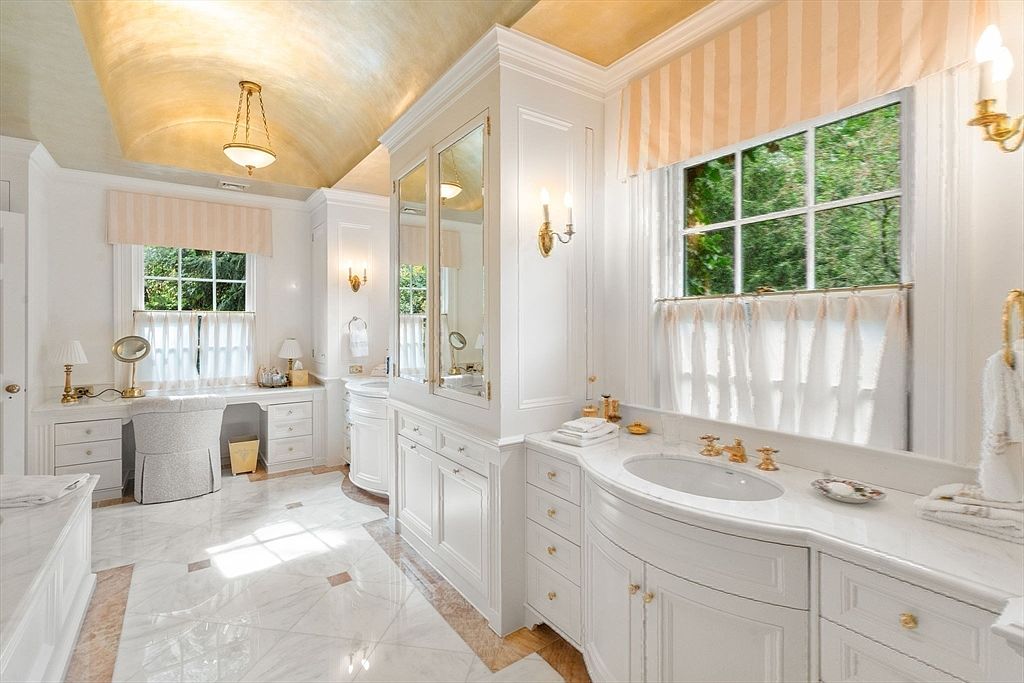 This elegant primary bathroom features a barrel-vaulted ceiling with a warm, metallic finish and a bright, airy aesthetic. The space includes a dual-vanity setup with white cabinetry, stone countertops, and a dedicated makeup vanity area with a matching chair. Large windows with soft, striped valances and sheer curtains allow natural light to fill the room, highlighting the polished stone flooring.