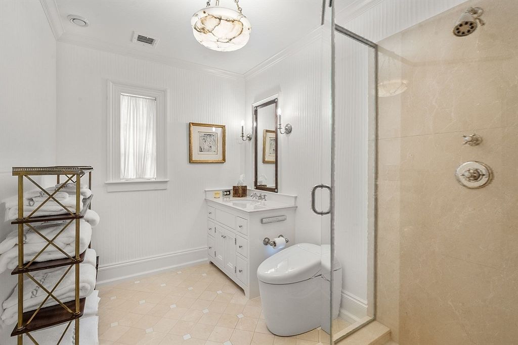 This bright and elegant bathroom features a white vanity with a stone countertop, a modern toilet, and a glass-enclosed shower with stone-tiled walls. A tall metal shelving unit holds neatly stacked white towels, while a decorative light fixture hangs from the ceiling. The room is finished with light-colored tile flooring and a window that allows natural light to fill the space.