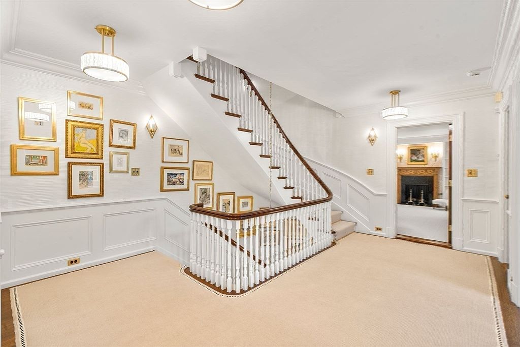 This elegant foyer features a grand staircase with a dark wood handrail and white balusters, set against a wall adorned with a gallery of framed artwork. The space is finished with crisp white wainscoting and neutral carpeting, creating a bright and sophisticated atmosphere. A doorway in the background provides a glimpse into an adjacent room featuring a fireplace, adding depth and a sense of flow to the home's layout.