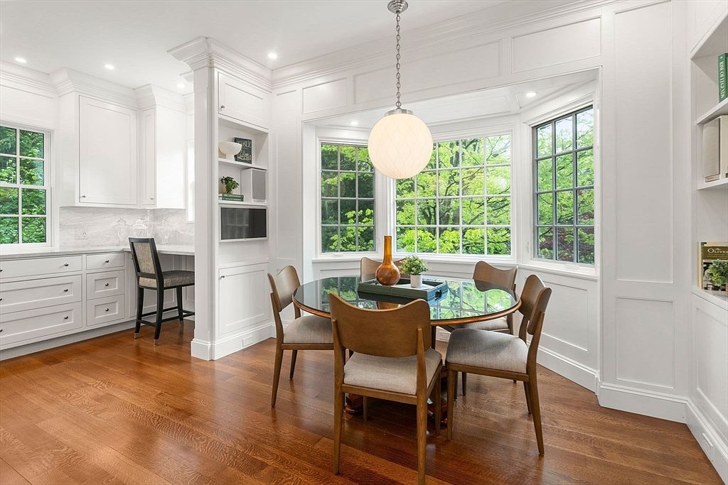 This bright and airy dining area features a round glass-top table surrounded by four modern wooden chairs with upholstered seats. A large, textured pendant light hangs above the table, centered in front of a bay window that offers a view of lush greenery outside. To the left, a built-in desk area with white cabinetry and a matching chair provides a functional workspace, while the room is finished with warm-toned wood flooring and crisp white trim.